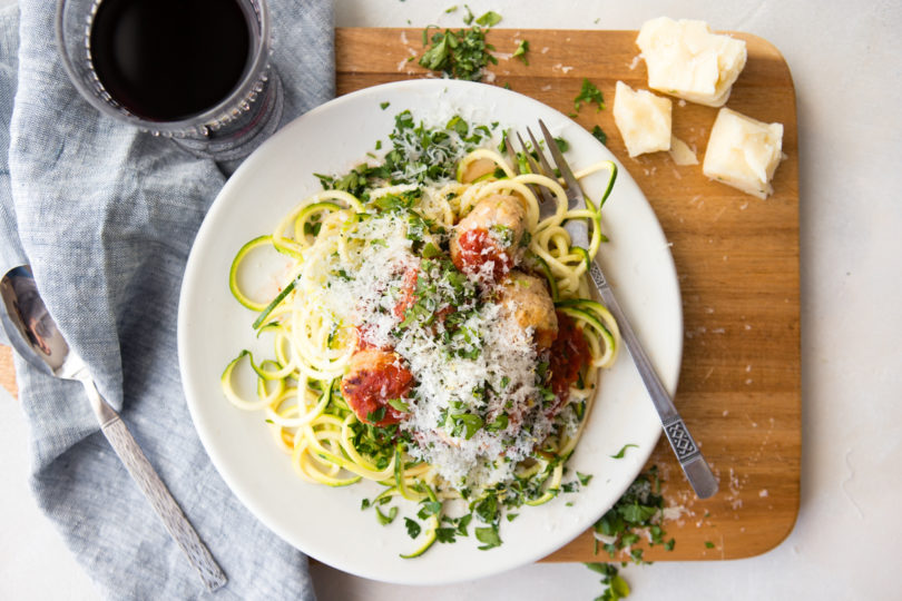 turkey meatballs and zucchini noodles and marinara sauce on a white plate set on a wooden board with a glass of red wine