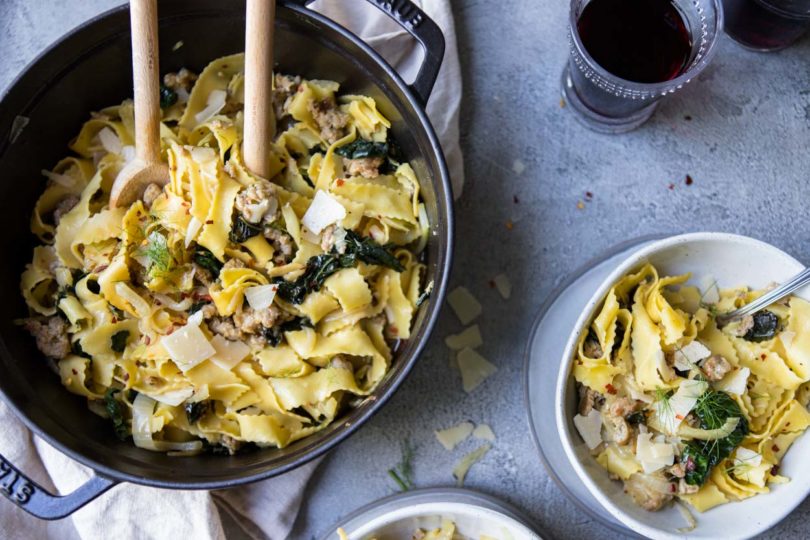 Wide egg noodles with sausage, kale and fennel in a black cast iron pot and on 2 gray bowls on a gray surface