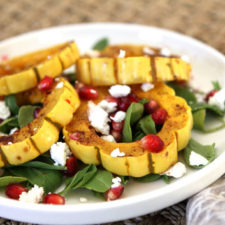 a white plate with roasted delicata squash, greens, pomegranate, and goat cheese