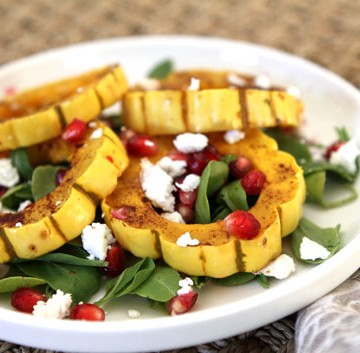 a white plate with roasted delicata squash, greens, pomegranate, and goat cheese