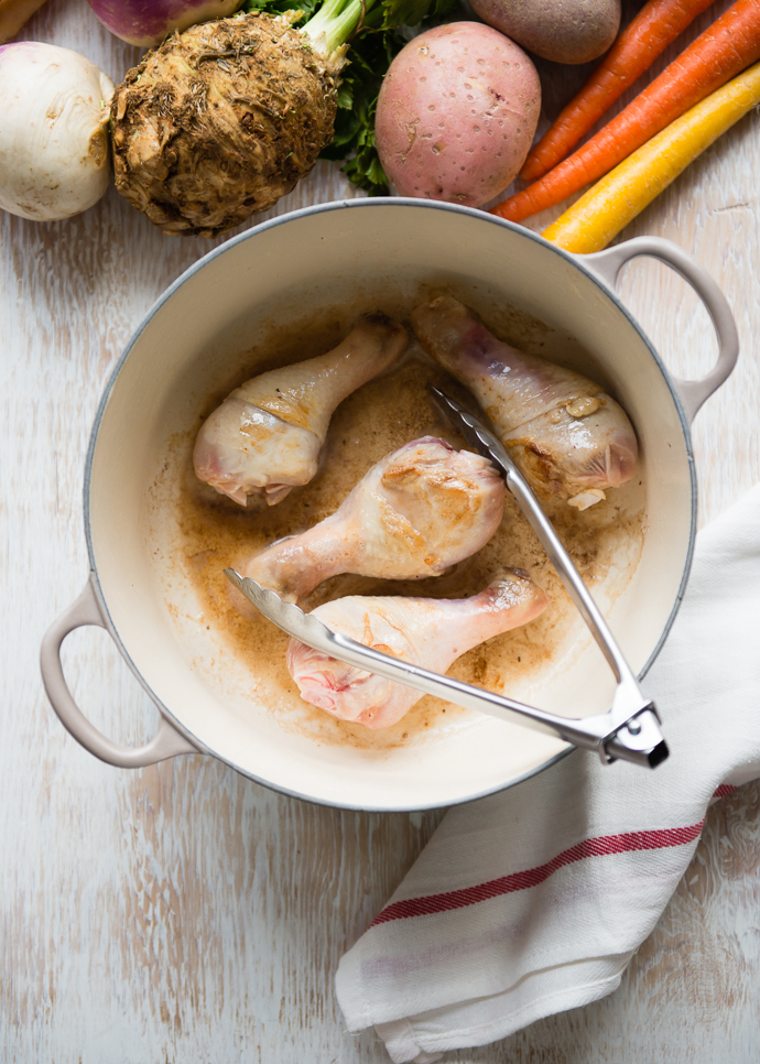 chicken pieces sautéing in a Dutch oven sitting next to potatoes and root vegetables 