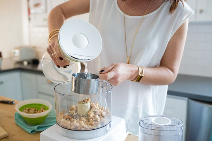 Pouring water into a measuring cup over a food processor filled with cashews