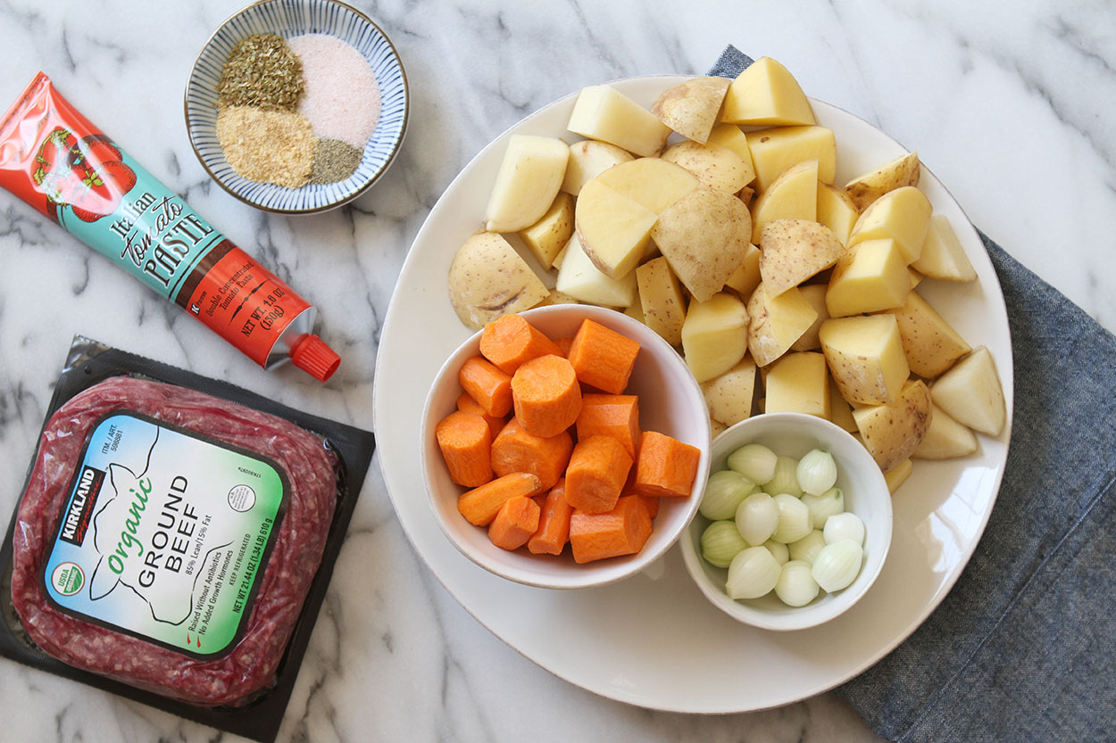 A white plate with potatoes, pearl onions, carrots. A pound of organic ground beef, tomato paste and a small bowl of herbs and spices.