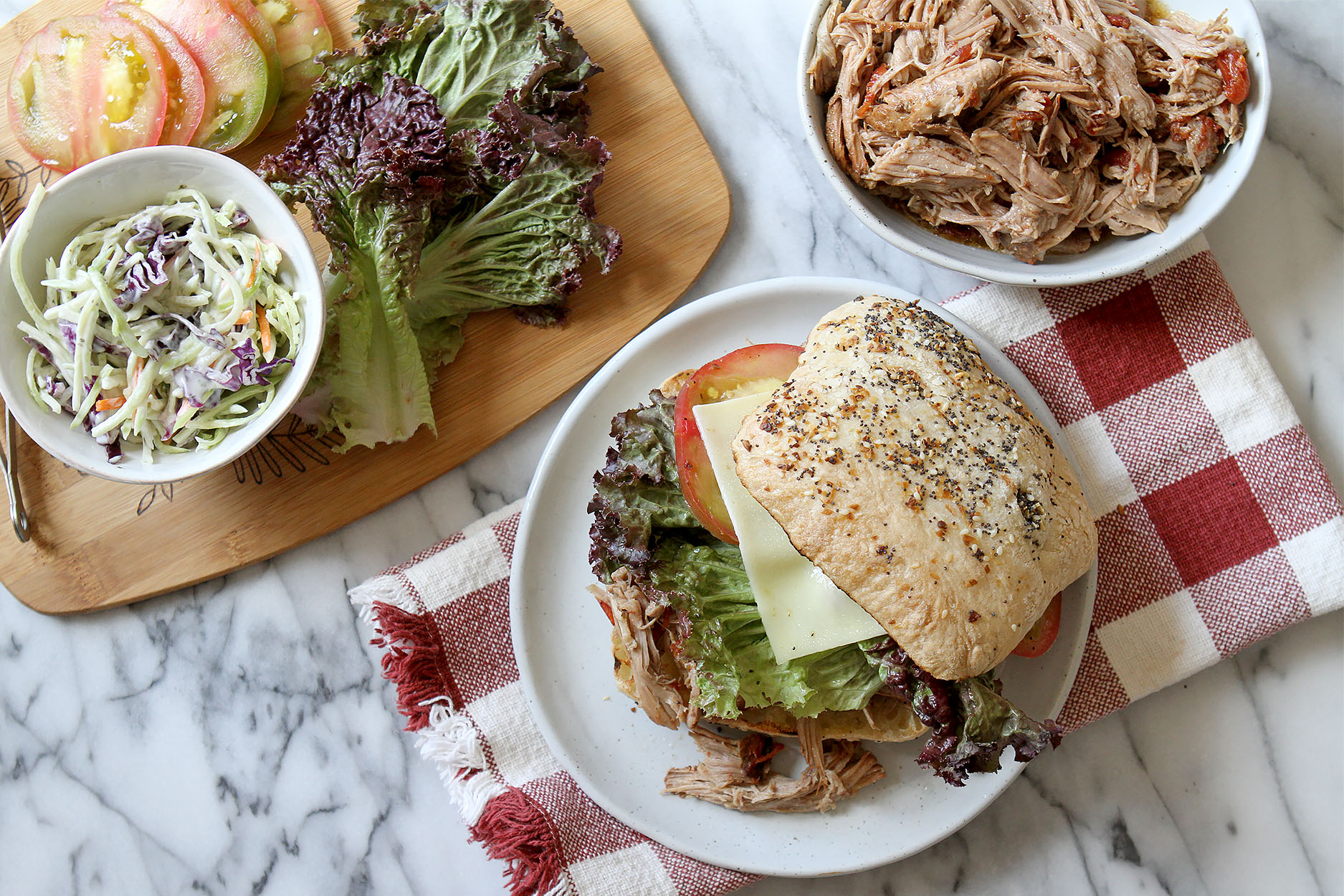 pulled pork sandwich on a white plate sitting on a red and white gingham napkin, a bowl of pulled pork to the side and a cutting board full of lettuce tomatoes and slaw.