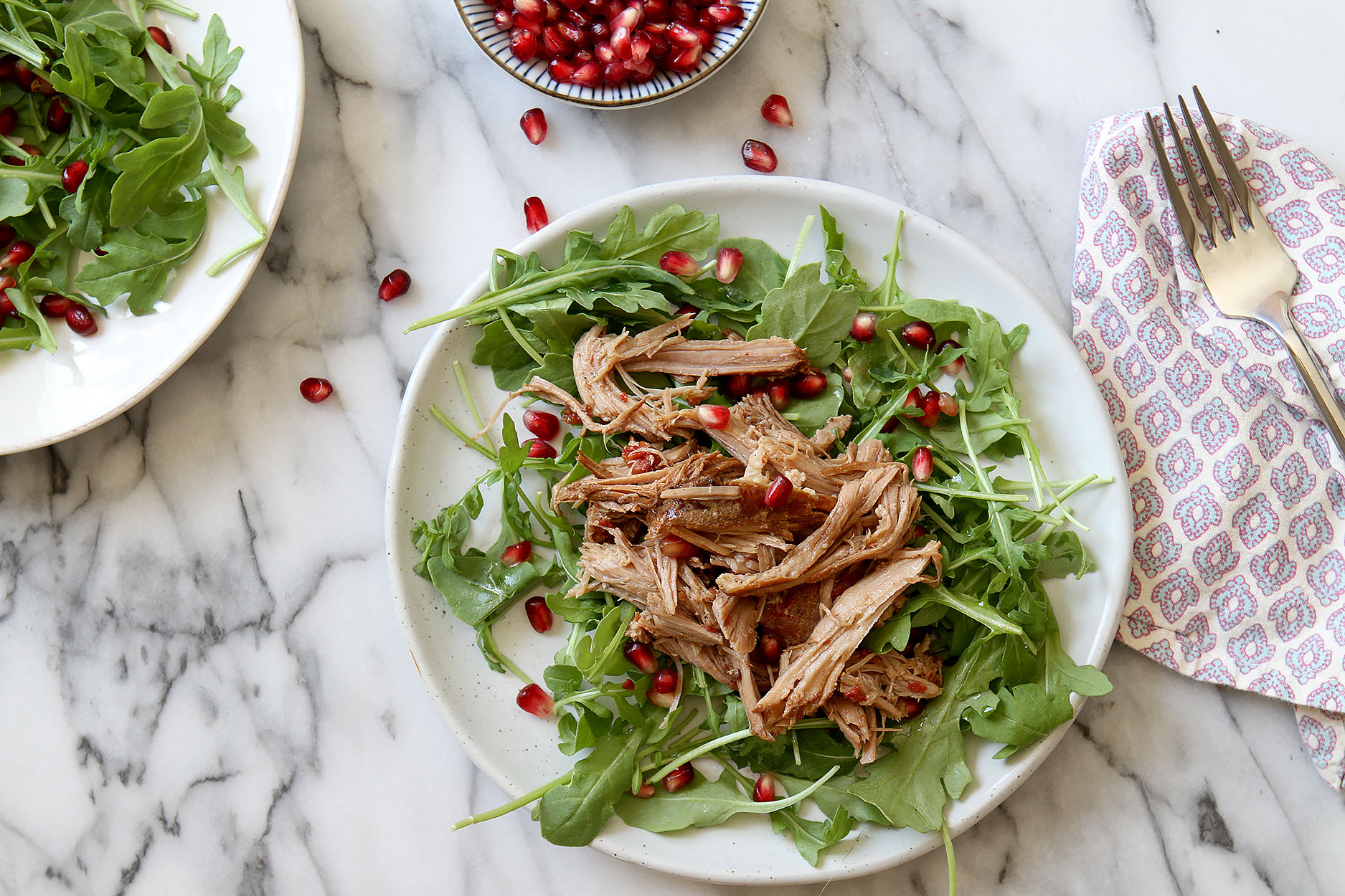 a white plate with arugula, pomegranate arils, and pulled pork, a napkin and fork to the side