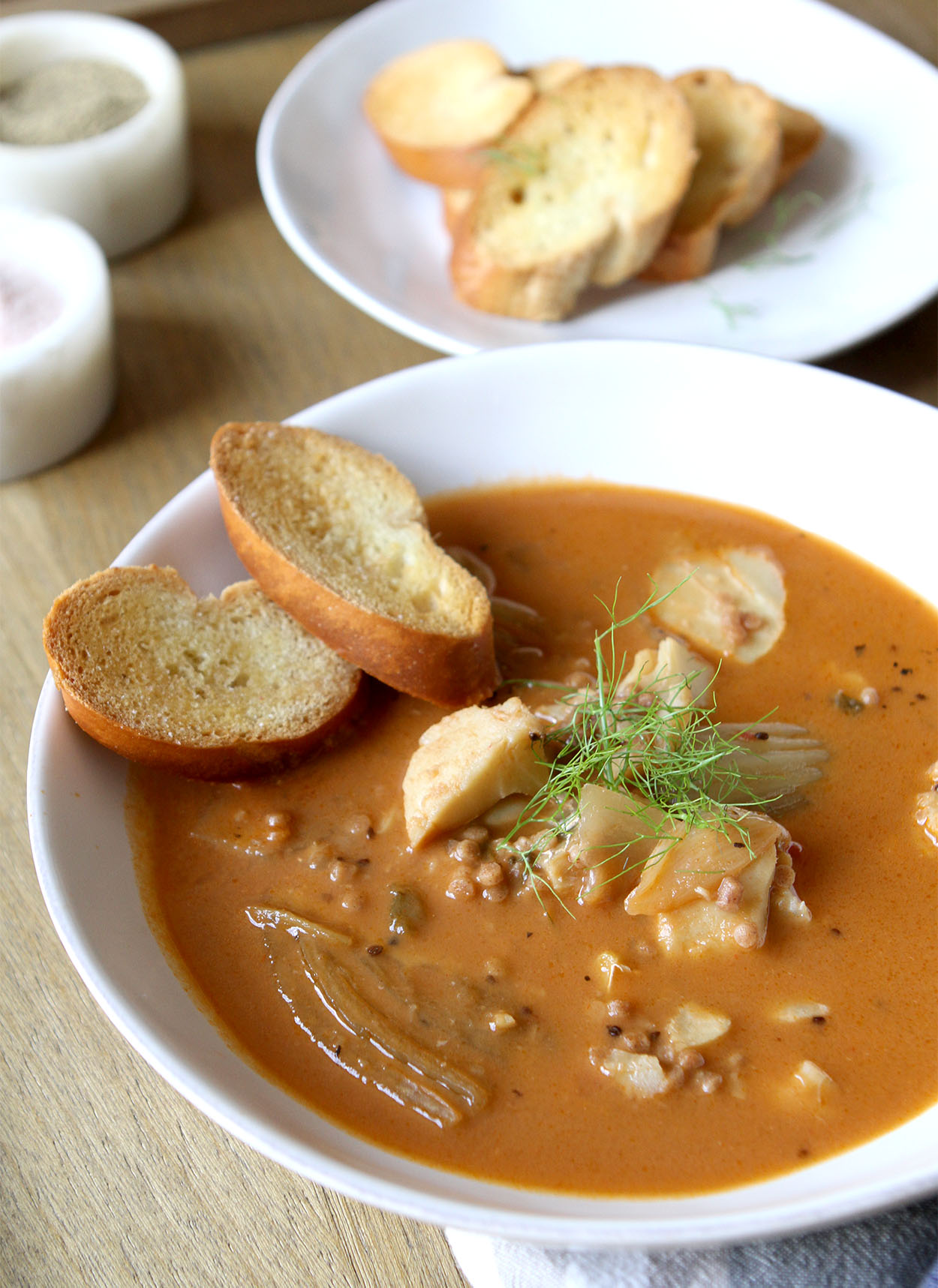 A white bowl full of fish stew, garnished with fennel frans and two crostinis