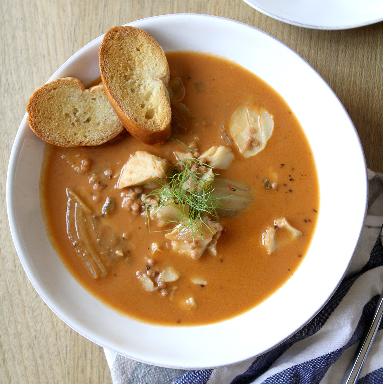 A white bowl of slow cooker fish stew, garnished with fennel frans and two crostini