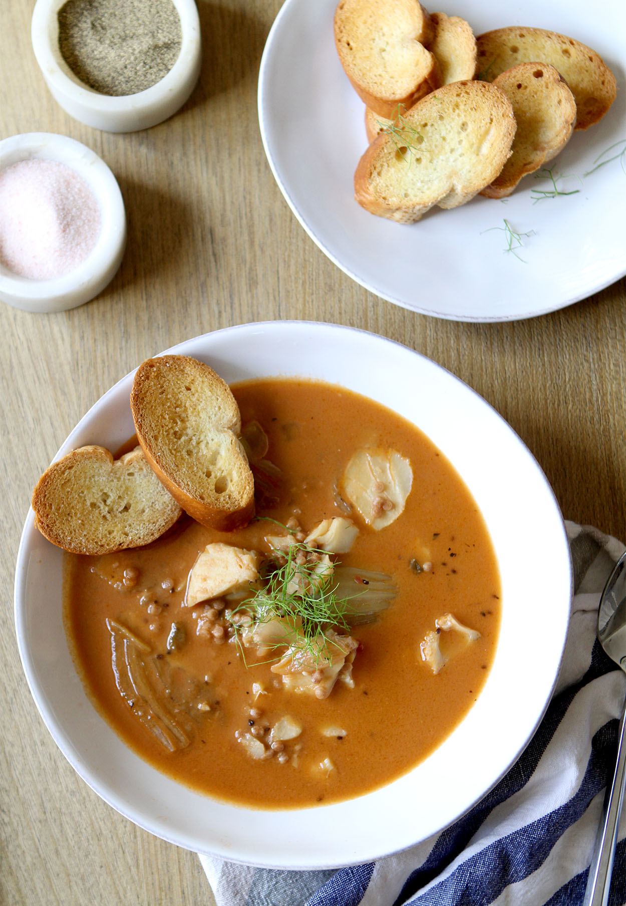 A white bowl full of slow cooker fish stew garnished with fennel frans and two crostini, a bowl of crostini and smaller bowls of salt and pepper behind