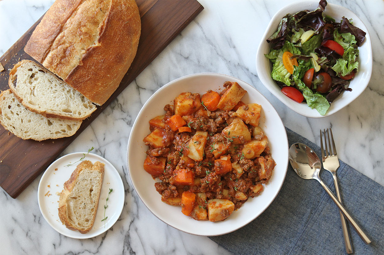 a white bowl of slow cooker beef stew, a loaf of bread on a wooden cutting board and a green salad in a white bowl. A fork and spoon to the side