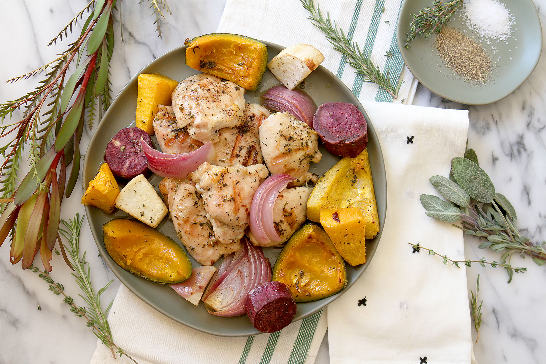 A round plate full of chicken and roasted root vegetables, herbs and greenery to the side