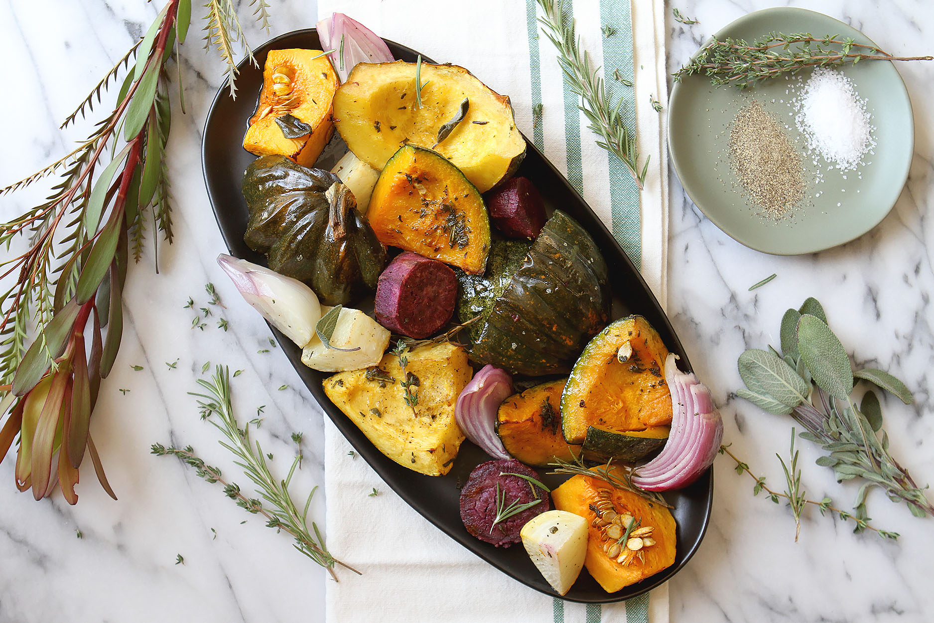 an oblong black plate full of roasted squash, onions, turnips, and beets. Herbs and greenery to the side