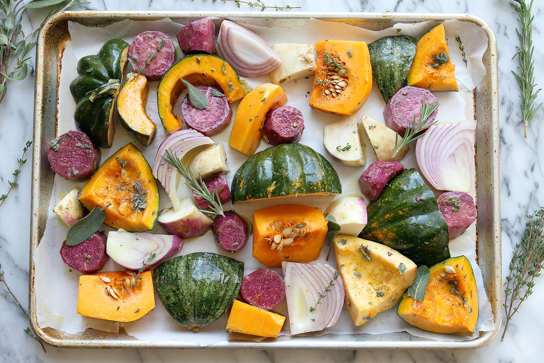 A sheet pan lined with parchment paper, full of squash, red onions beets, and turnips. Thyme, rosemary and sage sprinkled over the veggies