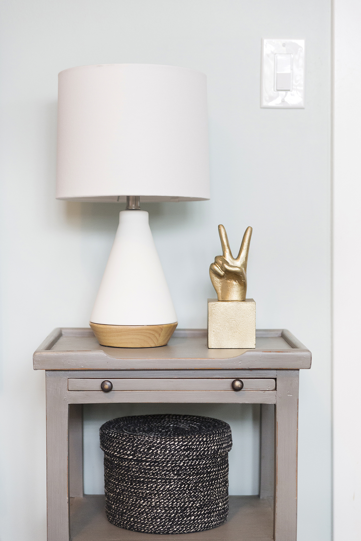 Night stand with a white lamp, gold peace hand and black basket