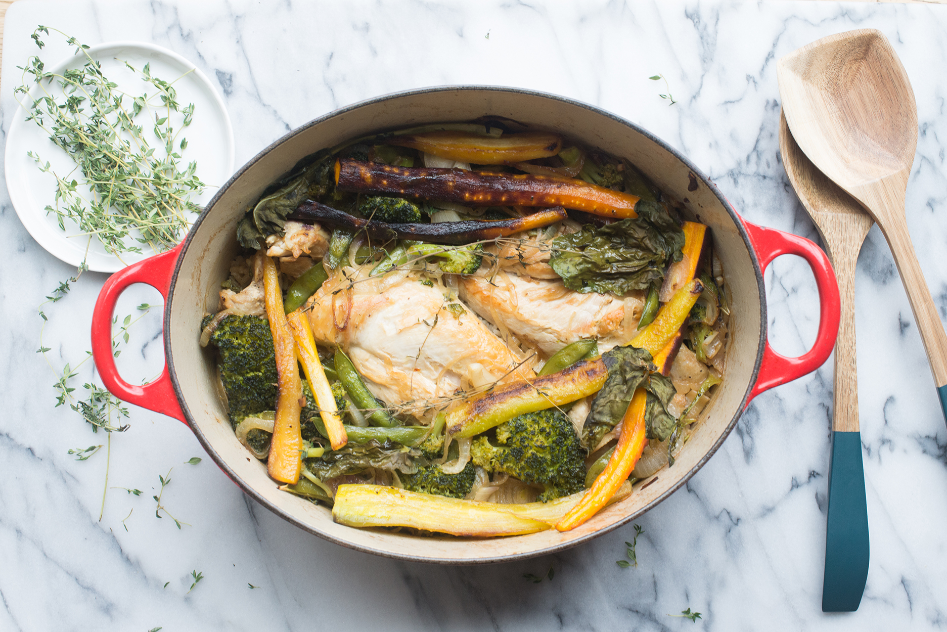 chicken and vegetables in a red dutch oven, with wooden spoons and a plate of thyme to the side