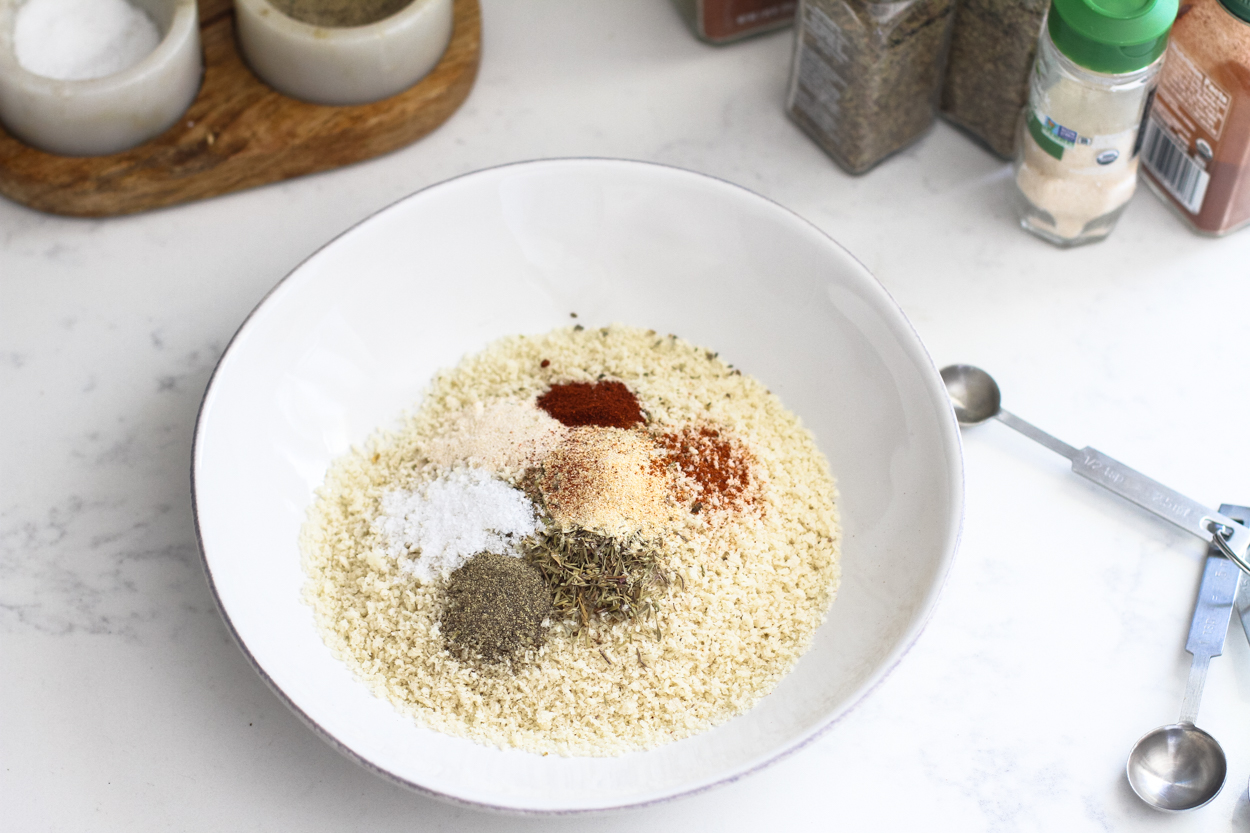 A white bowl full of bread crumbs and all the seasonings that go into the breading, measuring spoons to the side