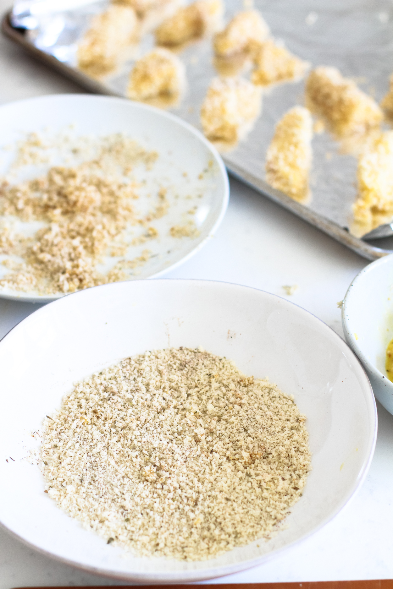 A white bowl full of breading, a baking pan of freshly breaded cod pieces in the background