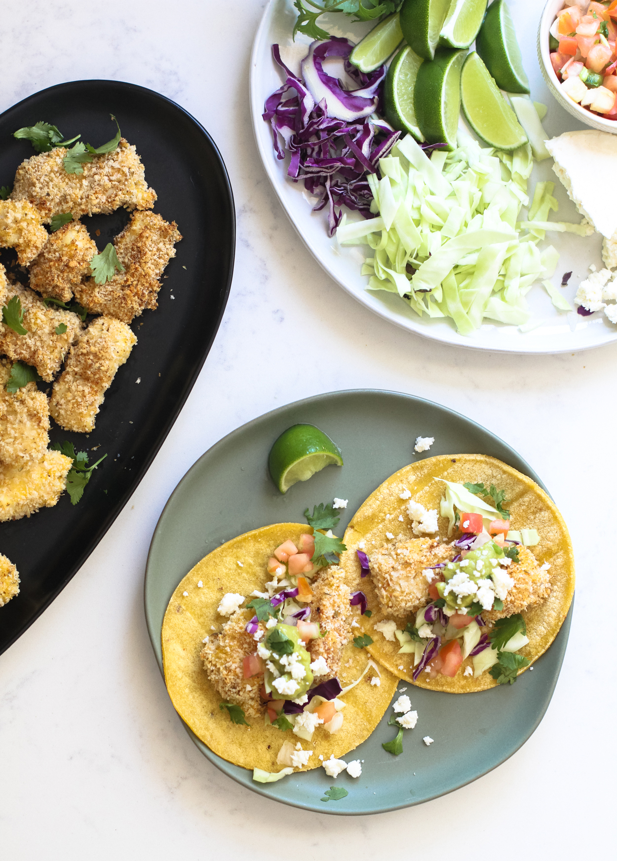 two crispy baked fish tacos on a round blue plate with a lime wedge to the side. A black plate with crispy cod pieces and a white plate with taco toppings in the background