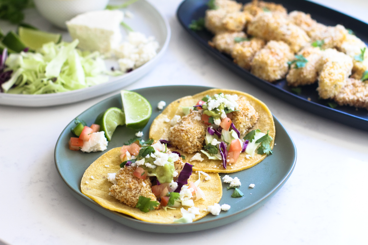 crispy baked fish tacos in tortillas on a blue plate with lime, tomatoes and queso fresco. A black plate with pieces of baked cod and a plate with lettuce and queso fresco in the background.