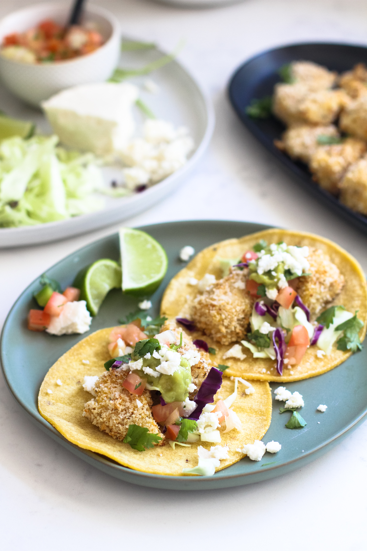 A blue plate with two baked fish tacos, tomatoes, queso fresco and limes to the side. Two plates of taco toppings and baked cod in the background