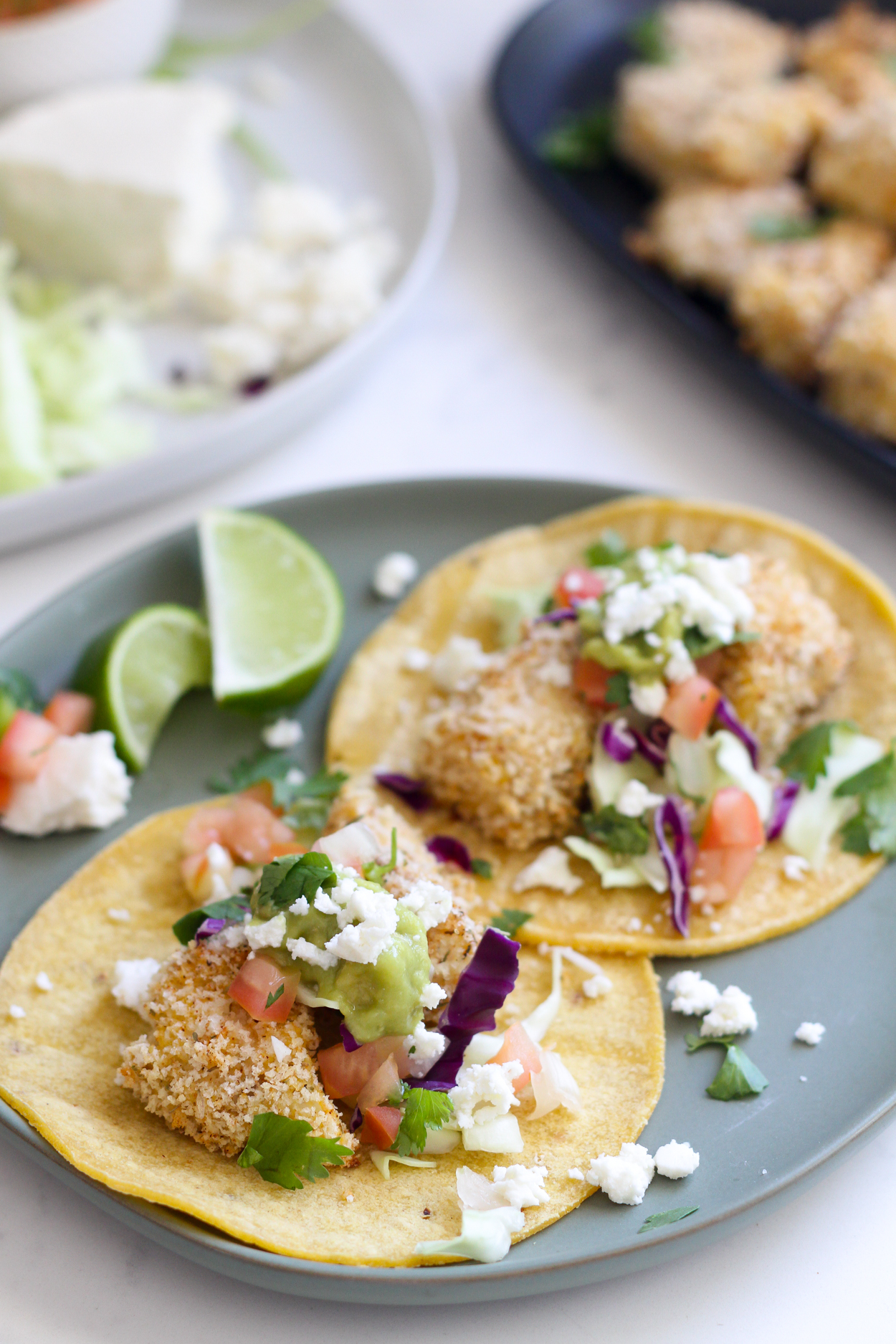 A blue plate with two crispy baked fish tacos topped with queso fresco tomatoes and lime wedges. Crispy baked cod pieces on a black plate in the background