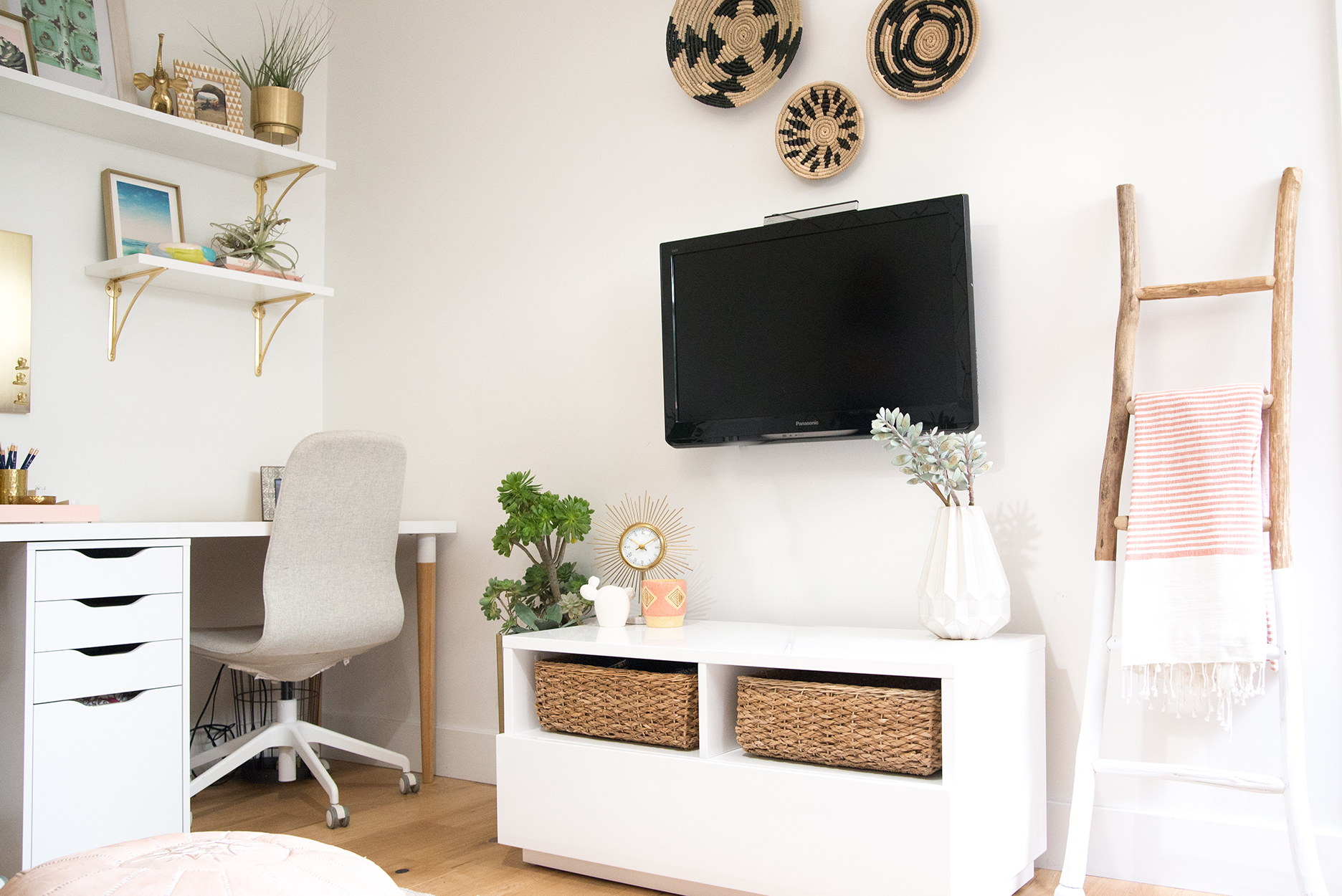 Office space with a white desk and grey chair with white and gold shelves above. A white TV console with baskets and a tv mounted on the wall above