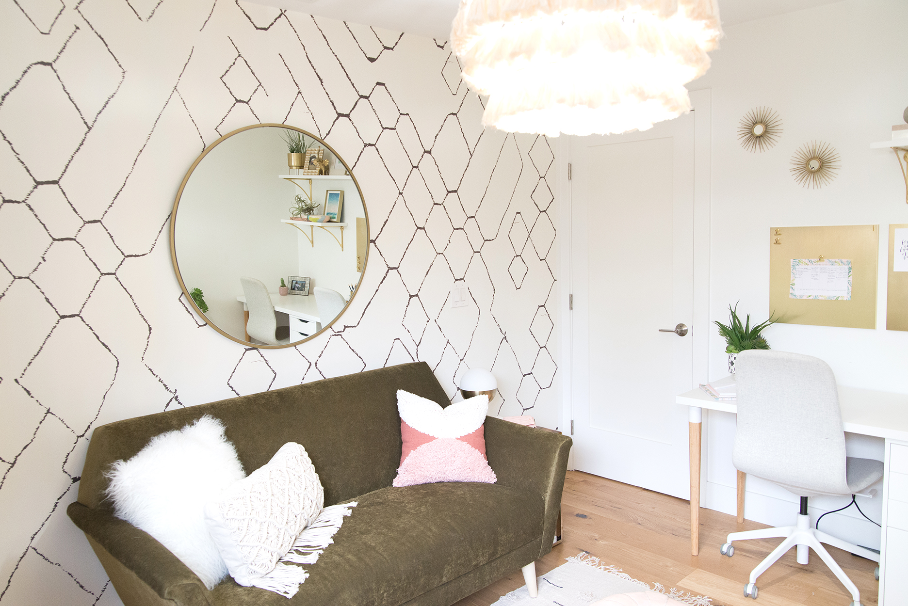 a geometric wallpapered wall with a round mirror, a small couch and a pink tassel chandelier