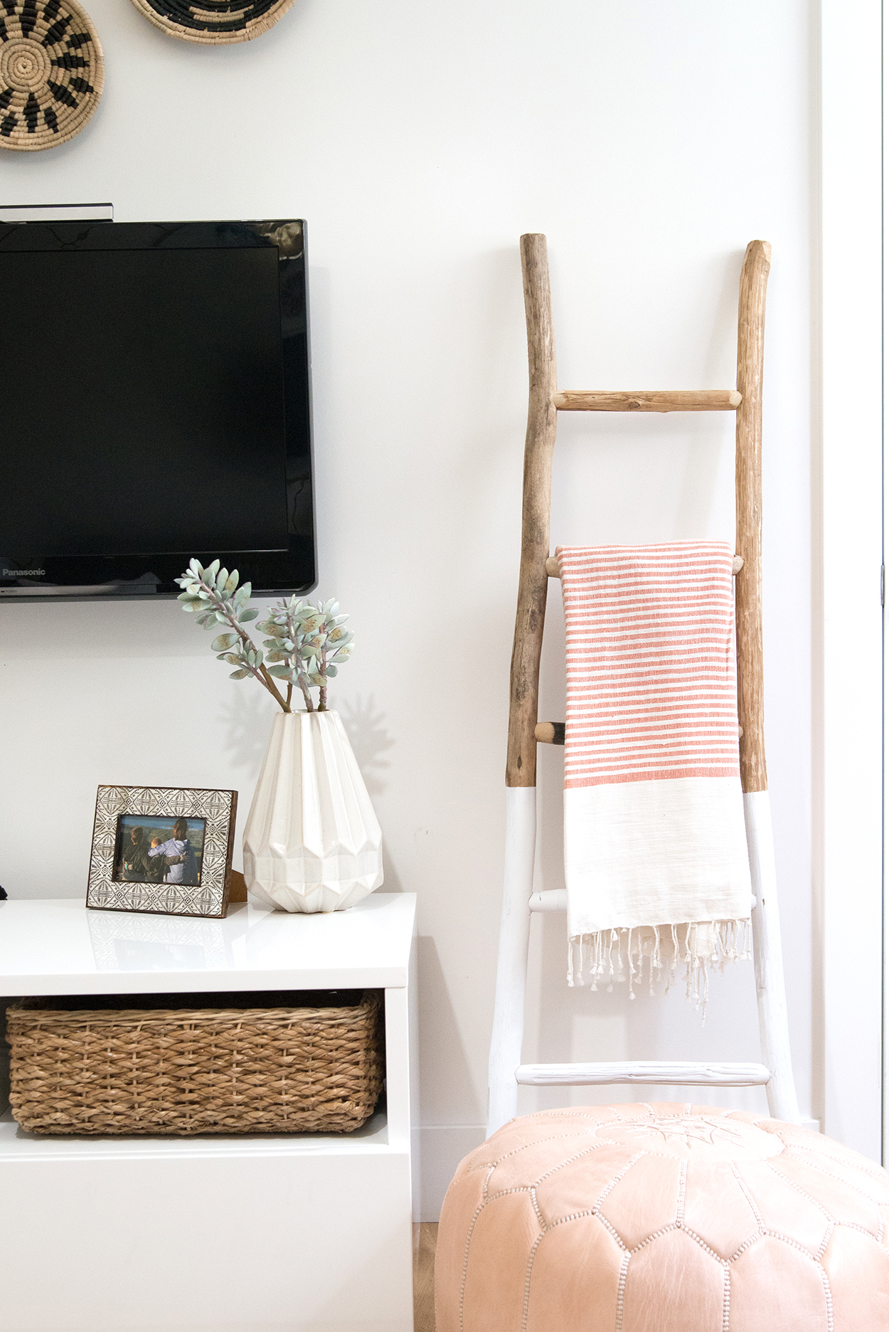 a wooden ladder with a stripped blanket thrown over a rung, a pink ottoman, and a white tv console 