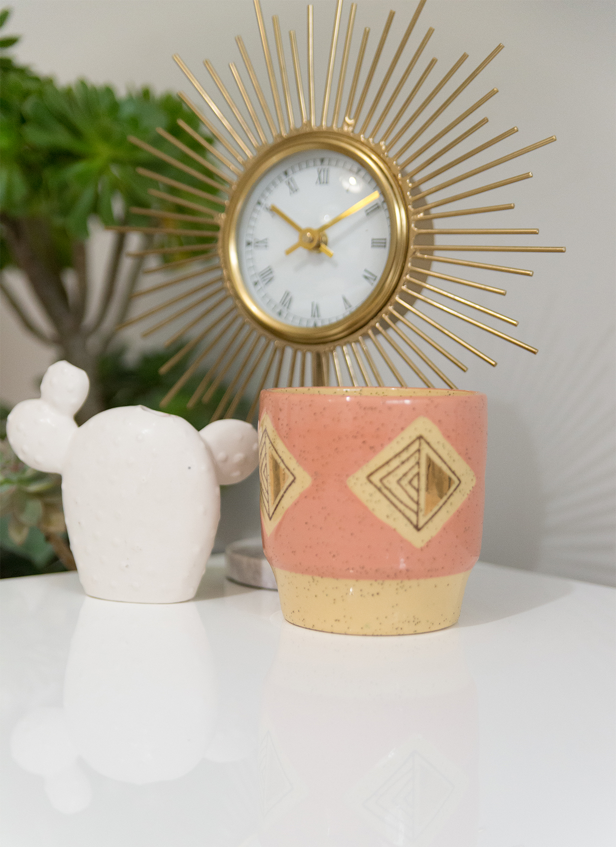 gold sunburst clock with a pink ceramic cup and a ceramic cactus sitting on the table