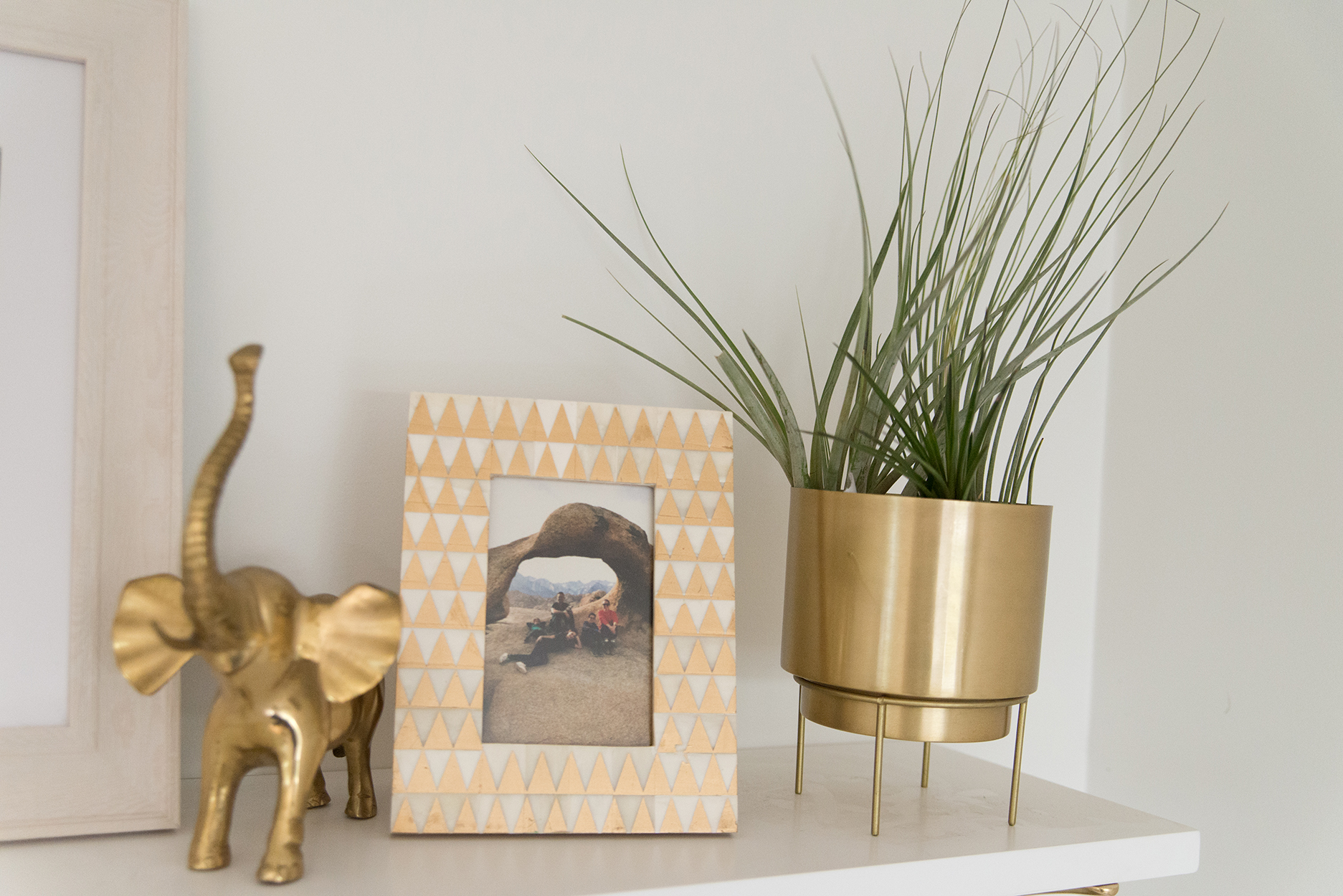 a white shelf with a plant potted in a gold pot, a dessert pic in a geometric printed frame and a gold elephant sculpture 