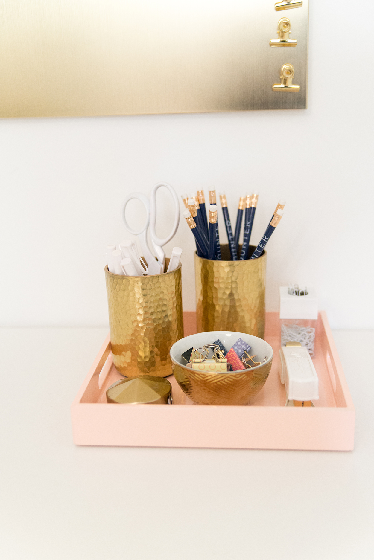 a pink tray holding a gold bowl of clips, and two gold cups holding pencils and scissors