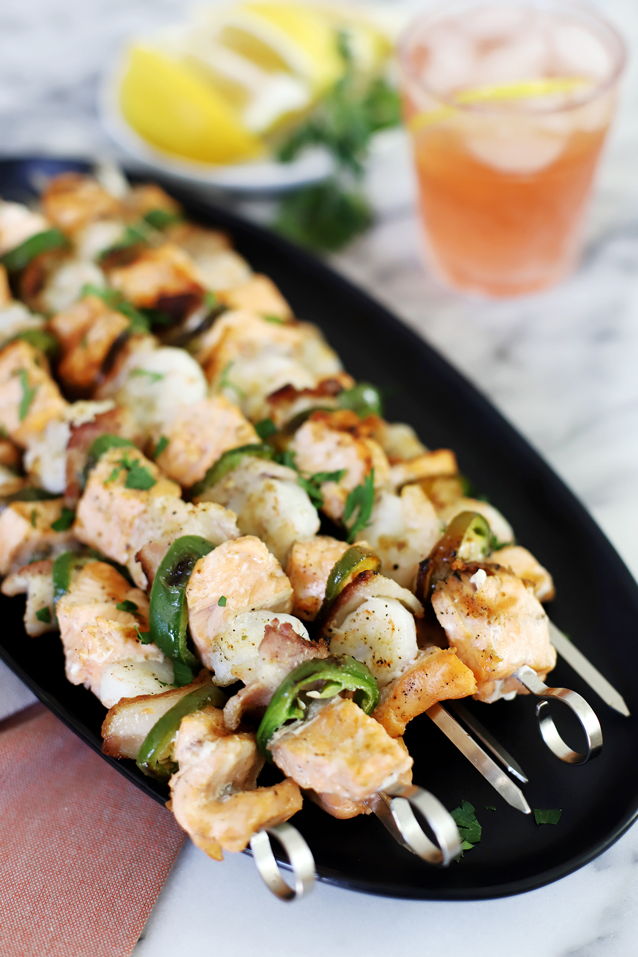 salmon and shrimp skewers on a black plate with lemons and a pink drink in the background