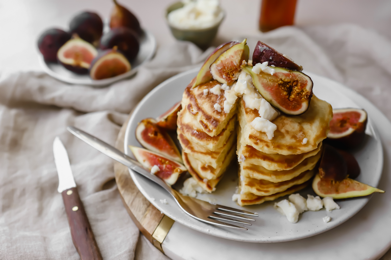 A stack of fluffy pancakes with a slice taken out, a knife to the side, and a plate of fresh figs and honey in the background