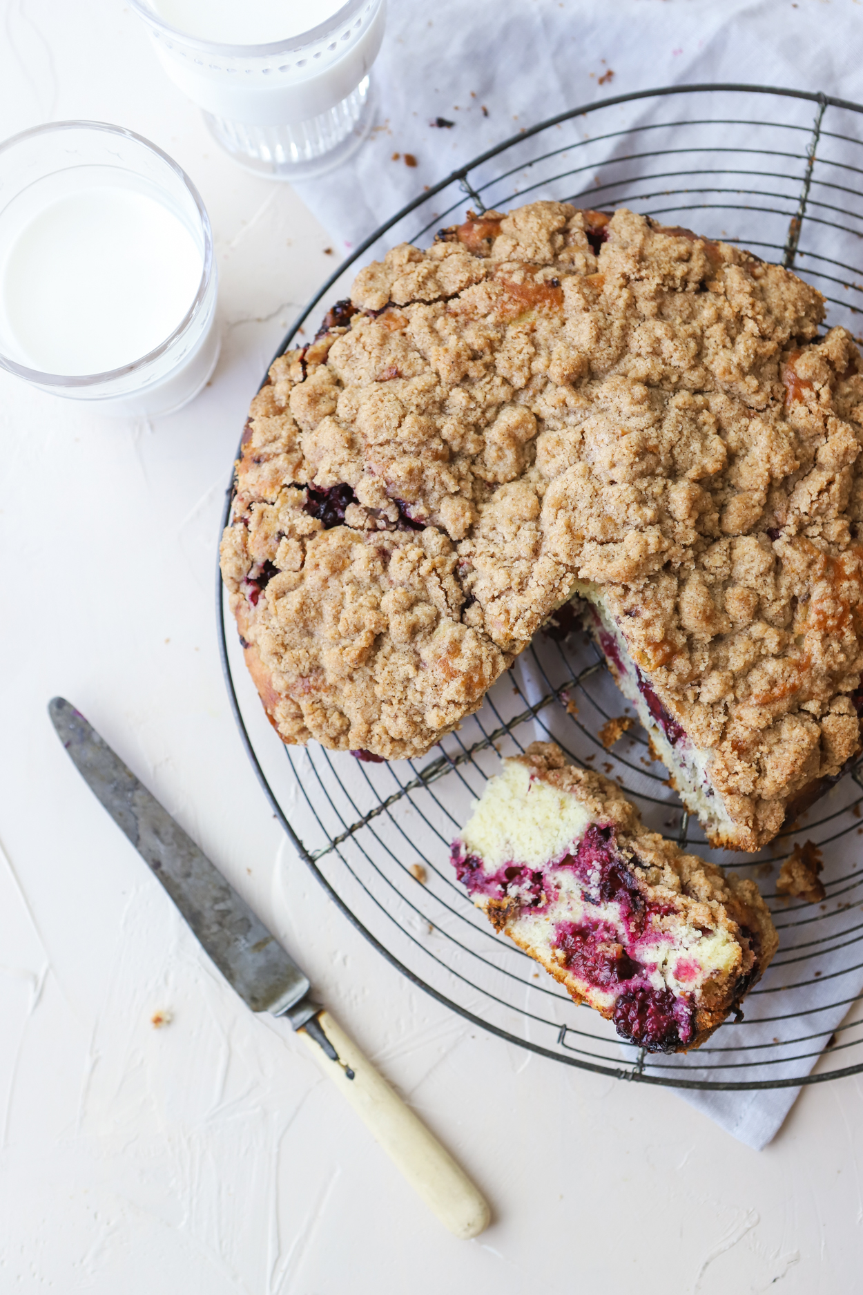 Blackberry crumble Cake on a wire cooling rack with a knife and a glass of milk on a white background