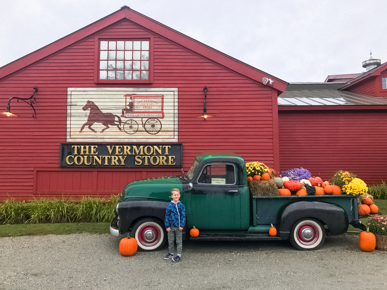 young boy standing in front of vintage green truck filled with pumpkins and mums in front of Vermont Country Store