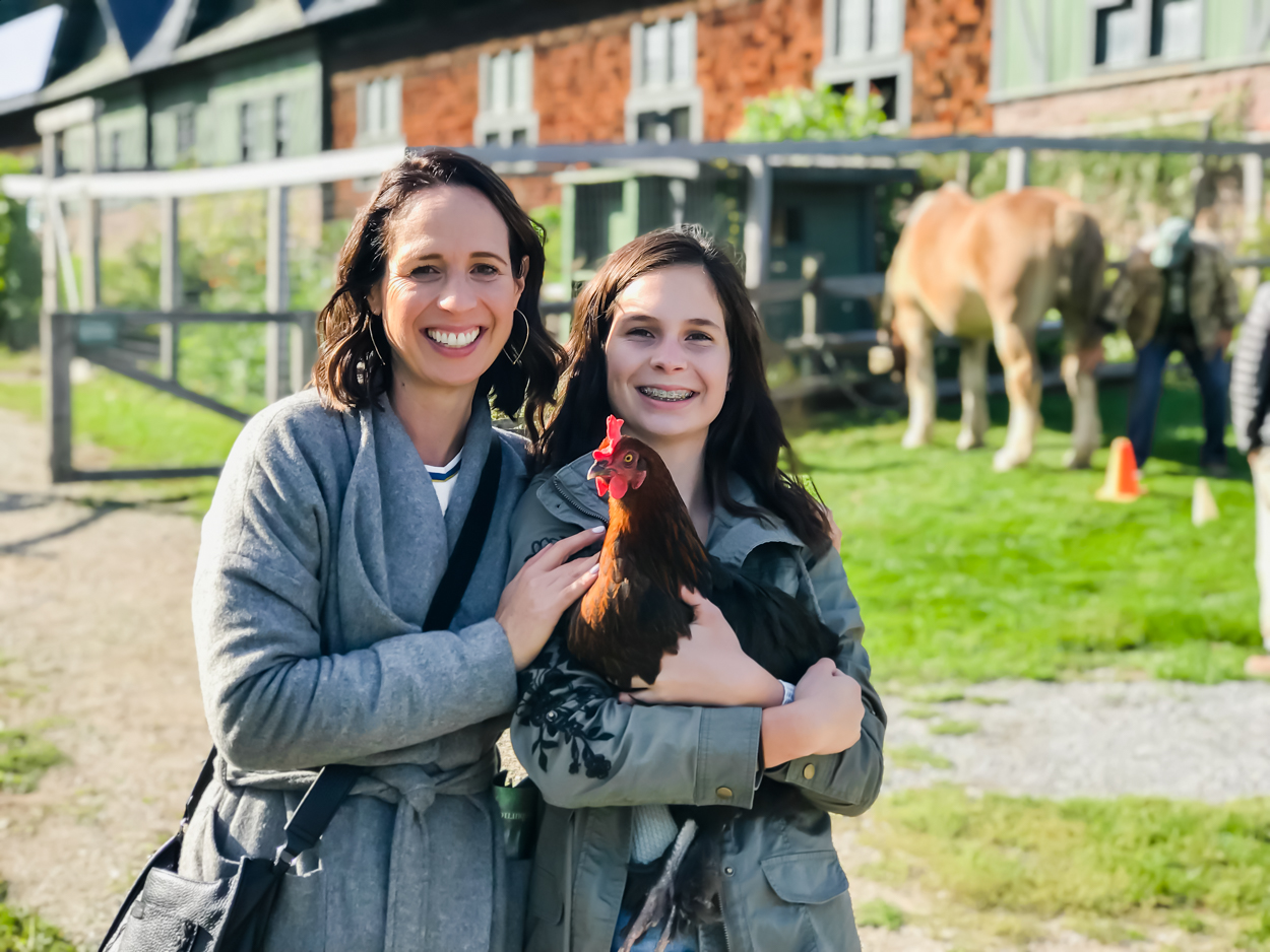 Mother and daughter wearing jackets standing in front of barn holding a chicken 