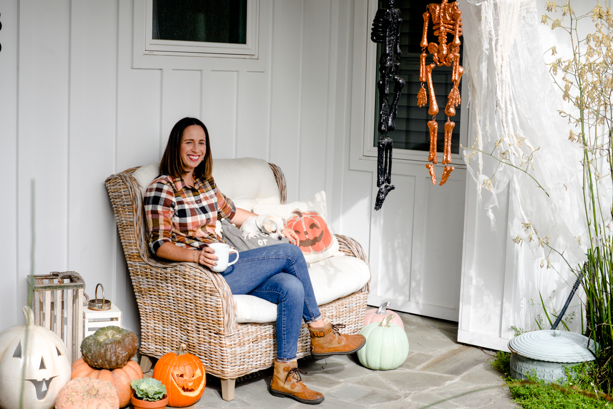 Woman with short brown hair wearing jeans and a plaid shirt sitting on a bench on a porch with a dog and Halloween decorations 