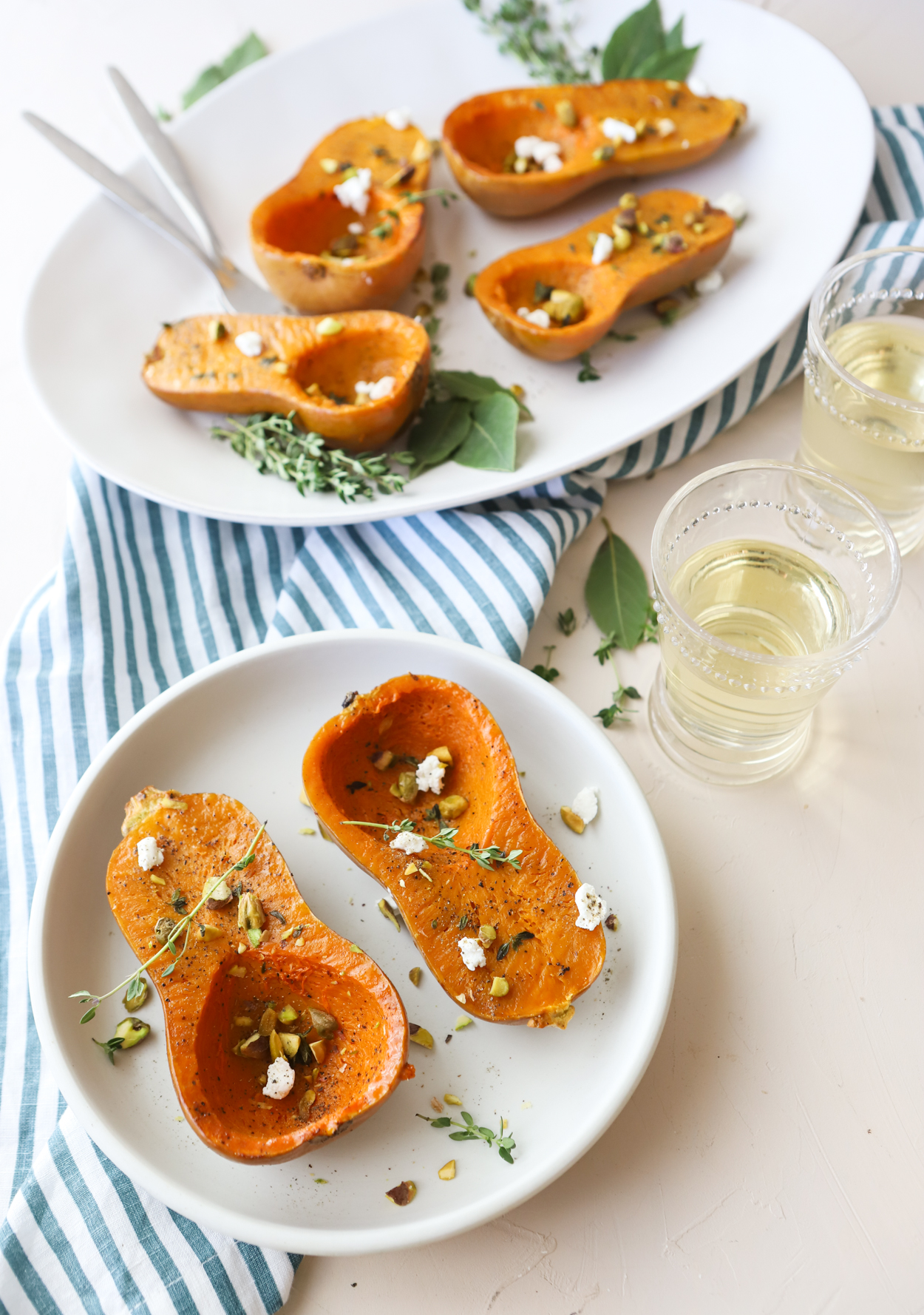 White platter and plate with squash cut in half sprinkled with goat cheese and pistachios and 2 glasses of white wine