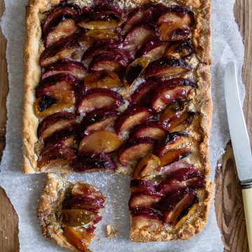 close up of a plum tart with a knife on top of parchment paper against a wooden backdrop