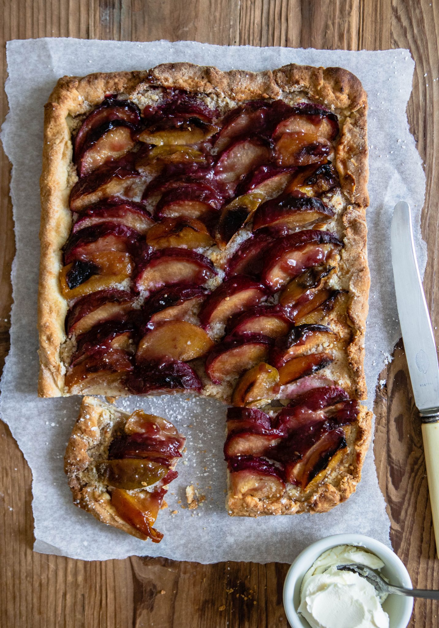 close up of a plum tart with a knife on top of parchment paper against a wooden backdrop