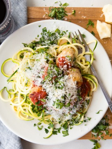healthy turkey meatballs and zucchini noodles and marinara sauce on a white plate with a fork set on a wooden board with a glass of red wine