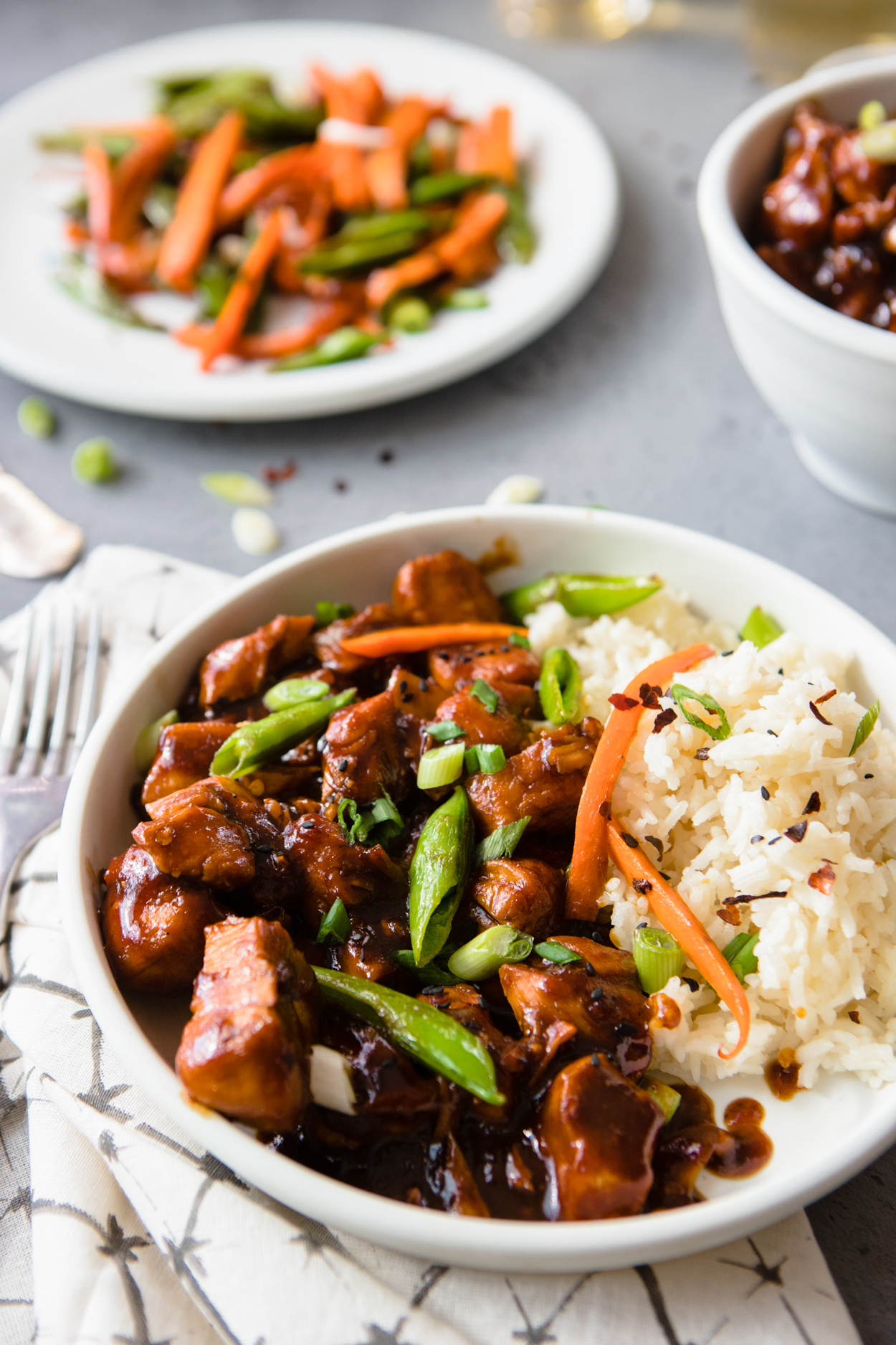 A white bowl full of General Tso's Chicken and rice garnished with scallions and carrots