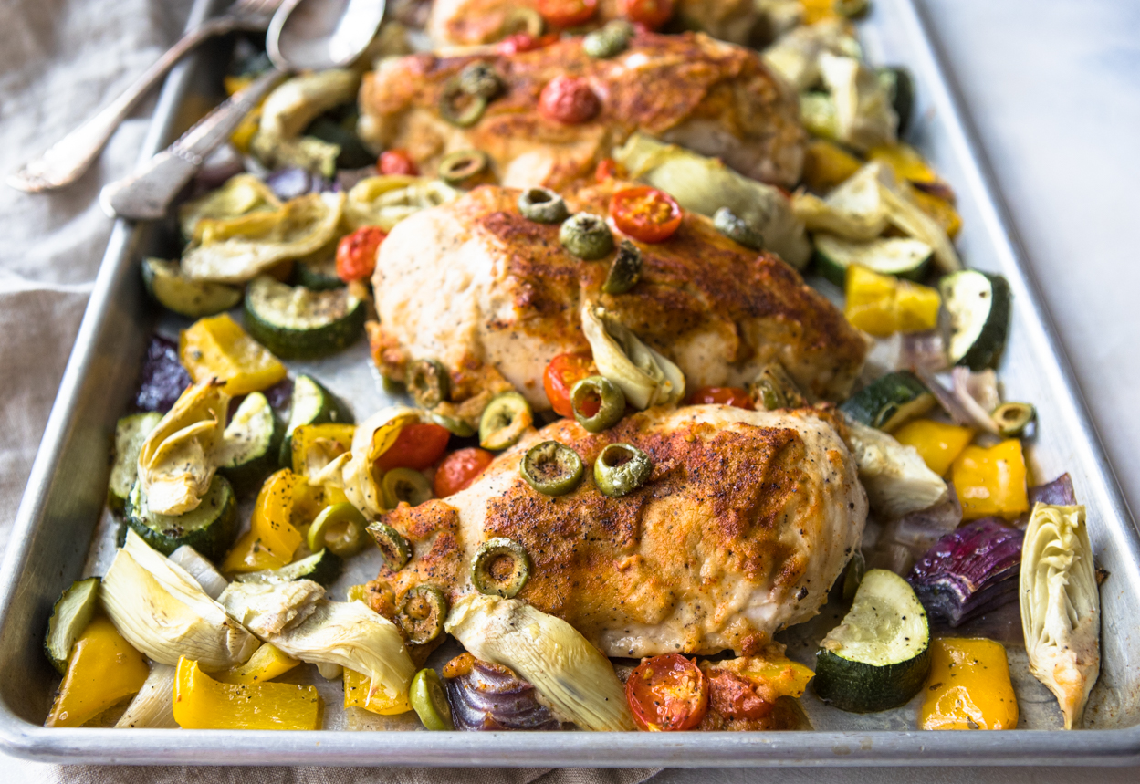 Hummus crusted chicken baked with vegetables, olives and <span style='background-color:none;'>artichoke hearts</span><span style='background-color:none;'> </span>on a rimmed <span style='background-color:none;'>baking sheet</span><span style='background-color:none;'> </span>with fork and spoon