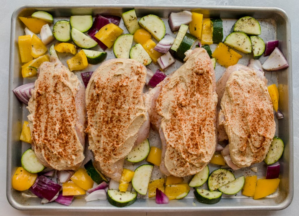 cut up vegetables and chicken breasts with hummus spread on them nestled on a rimmed baking sheet