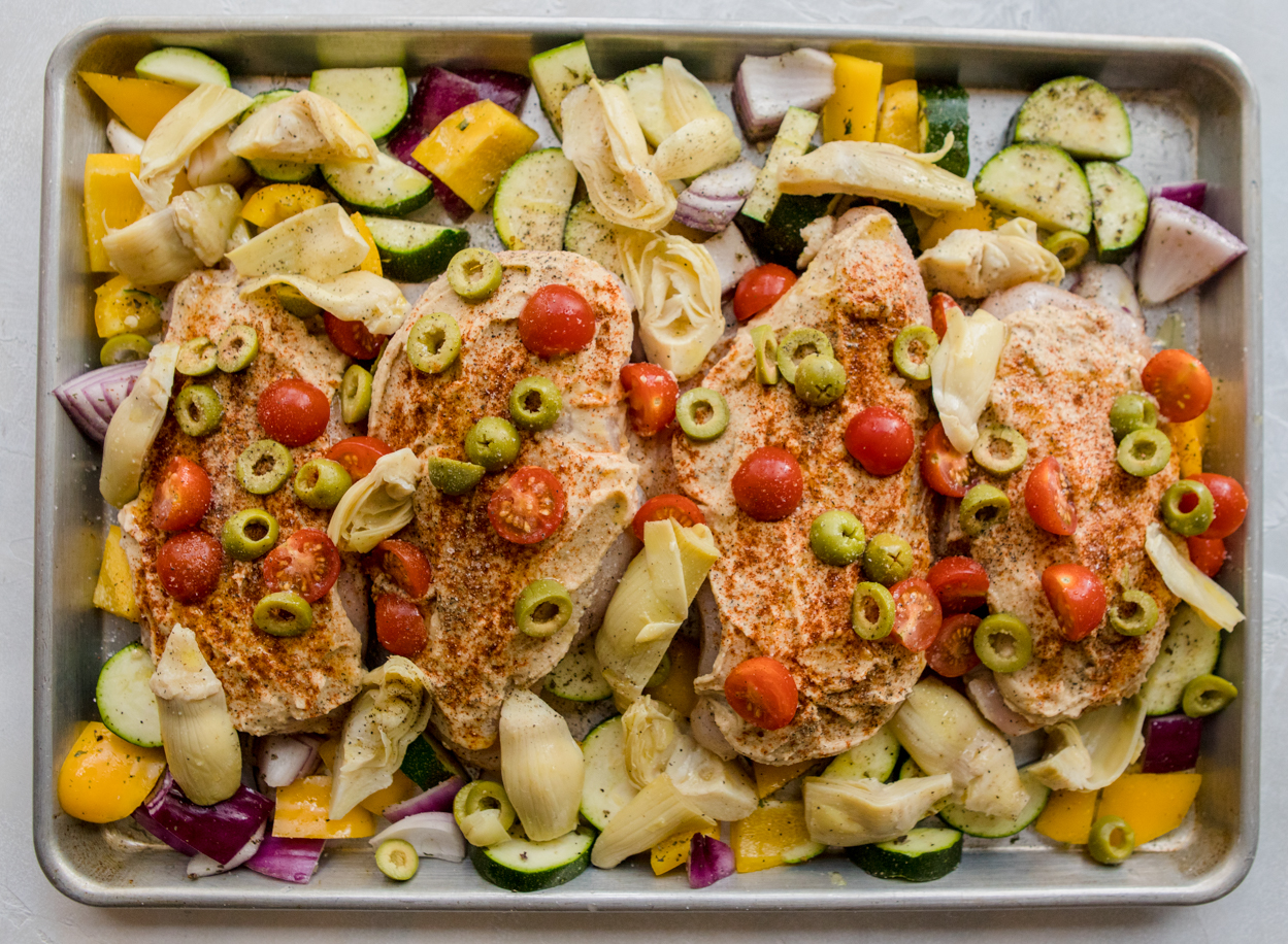 Cut up vegetables and chicken spread with hummus and green olives and tomatoes and artichokes on a baking sheet 