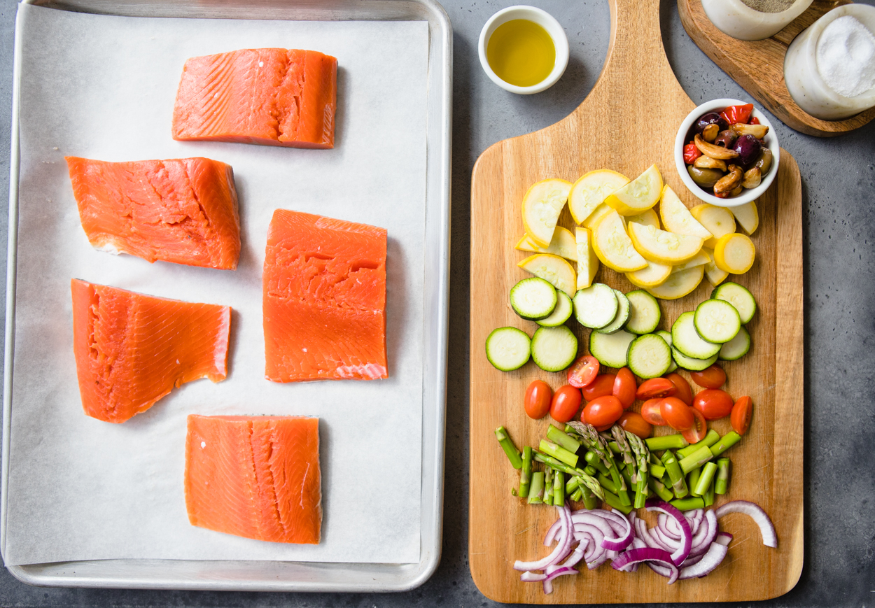 Sheet pan with parchement paper and salmon, veggies on a cutting board to the side