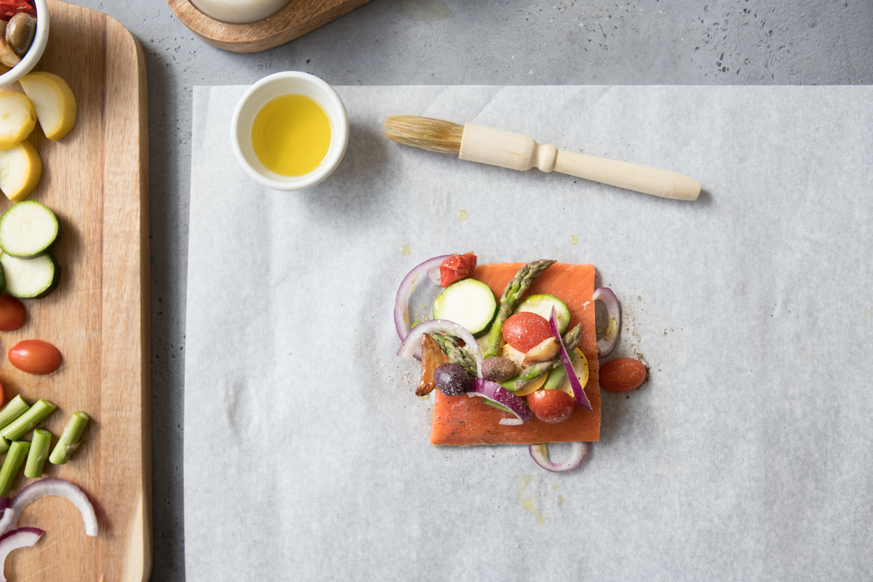 fresh salmon topped with veggies on parchment paper, olive oil to the side