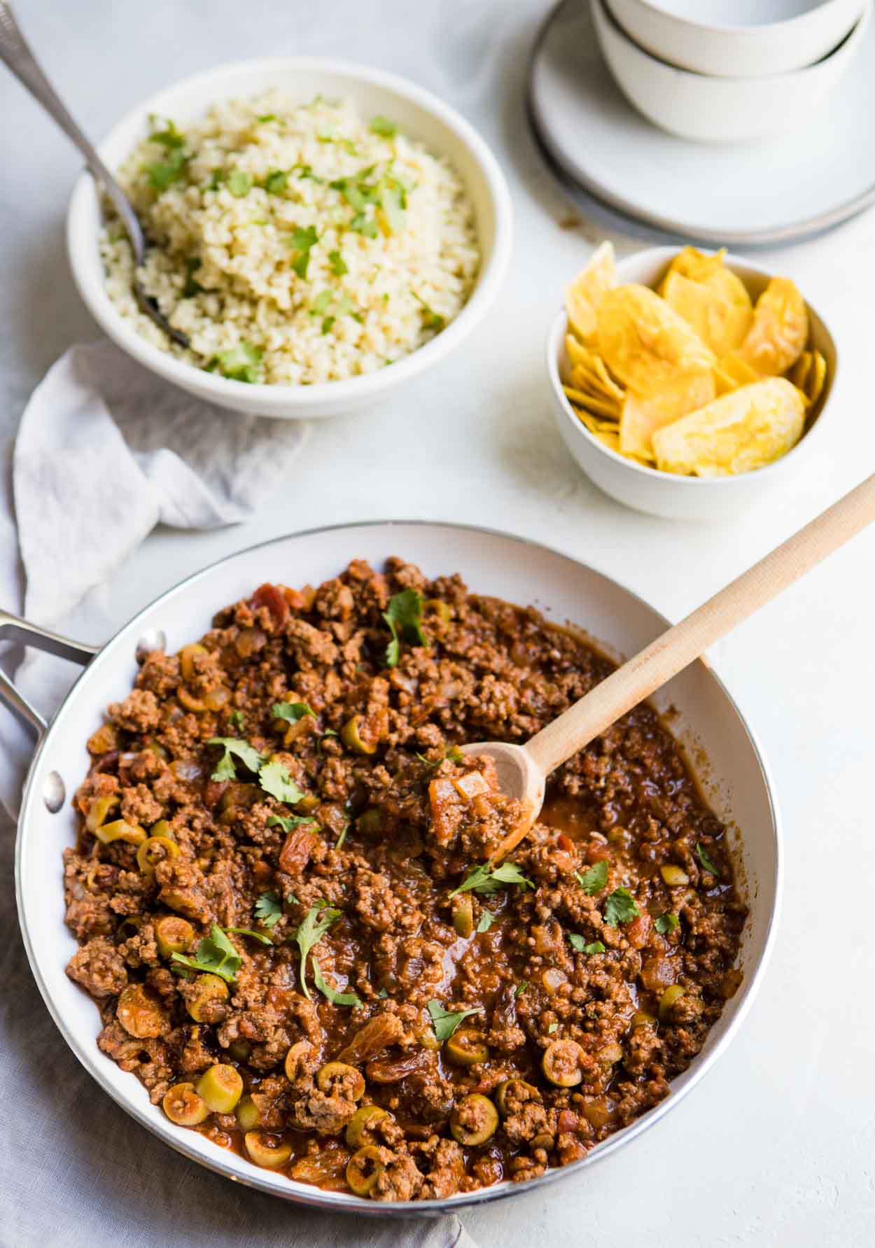 Ground beef picadillo in a white saute pan with a bowl of cauliflower rice and a bowl of plantain chips