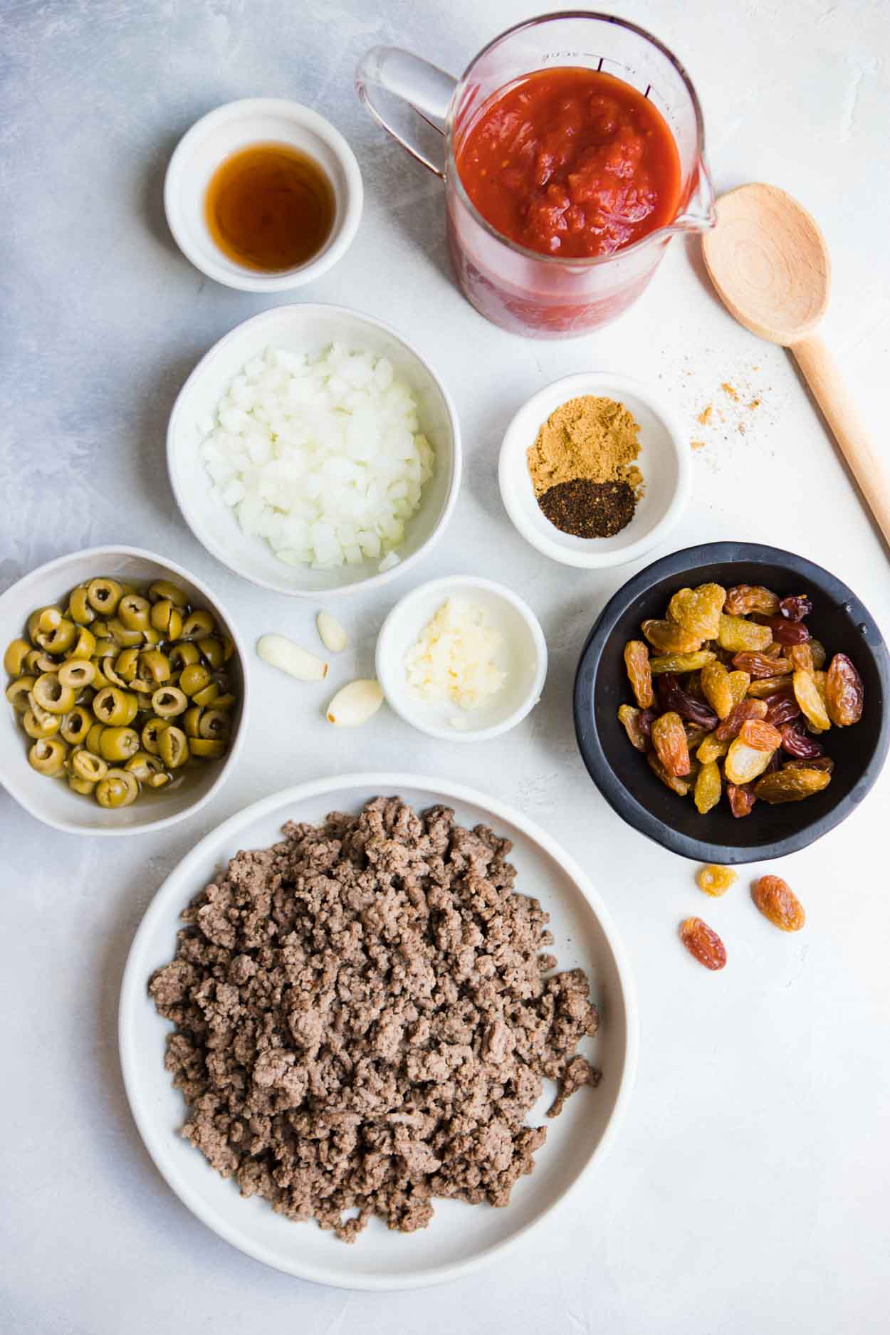 ground beef, raisins, green olives, onions, garlic and spices in a white bowl and tomato sauce in a glass cylinder