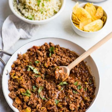 Ground beef picadillo in a white saute pan with a bowl of cauliflower rice and a bowl of plantain chips