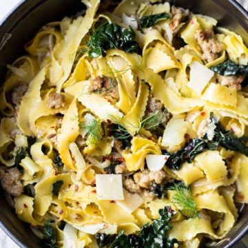 Wide egg noodles with sausage, kale and fennel in a black cast iron pot on a gray surface